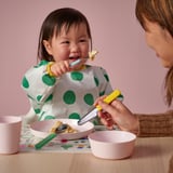 Child eats with colorful knife and fork, assisted by adult. Wearing polka dot apron, table set with bowls and mug. Bright, cheerful milestone.