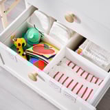 Open white nursery drawer unit with colorful items inside, including a yellow giraffe plush and folded red-striped towels.