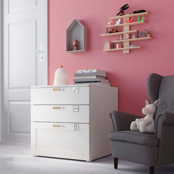 Childs room with pink walls, white storage unit, gray armchair, and shelves with toys.