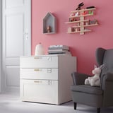 Childs room with pink walls, white storage unit, gray armchair, and shelves with toys.