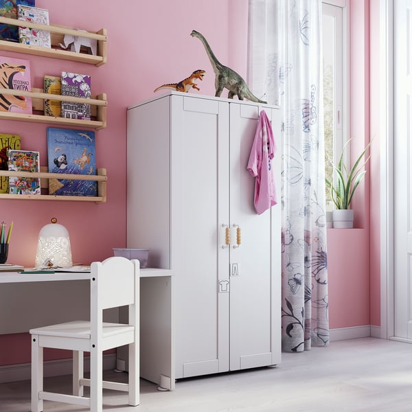 A kids room with pink walls, a white wardrobe with dinosaur toys, shelves full of colorful books, and a white desk with a chair.