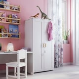 A kids room with pink walls, a white wardrobe with dinosaur toys, shelves full of colorful books, and a white desk with a chair.