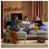 Three women relax on a sofa in a cozy living room. One works on a laptop, another drinks coffee, and the third reads.