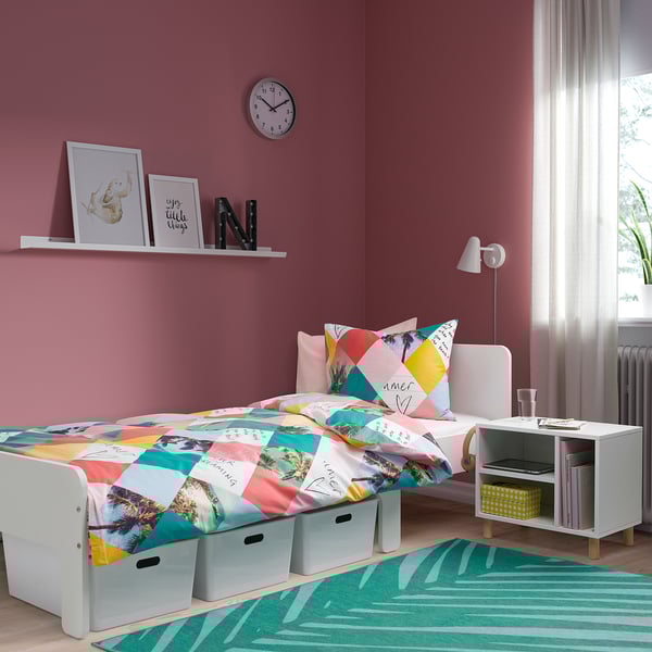 Bright pink kids room with white bedside table, yellow box, books, colorful bedspread, and striped green rug.