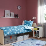 Bedroom with SMUSSLA white bedside table, floral rug, dinosaur-themed bedding, books, and decor on a pink wall.