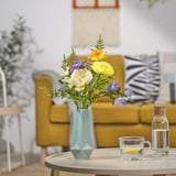Artificial flowers in mint green vase on wooden table with clear mug and carafe, yellow couch, and potted plant in background.