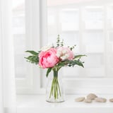 A clear glass vase holds a vibrant, life-like bouquet of pink flowers with white accents, displayed on a white window sill.
