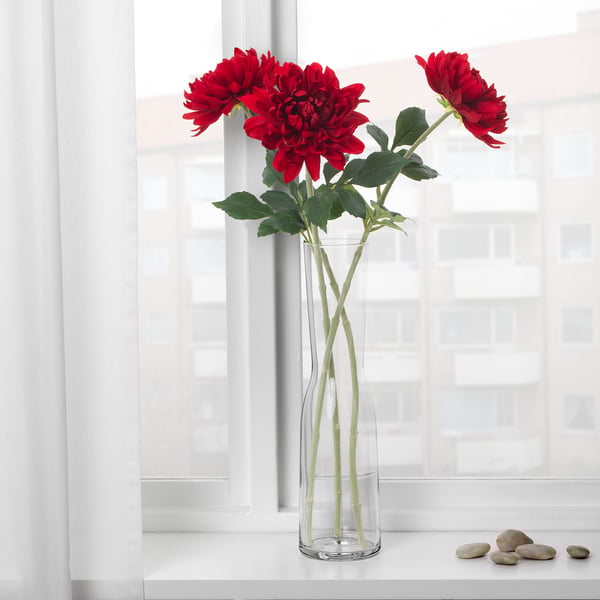 A clear vase with red artificial flowers on a windowsill. The flowers have green stems and leaves.