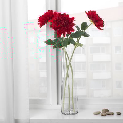 A clear vase with red artificial flowers on a windowsill. The flowers have green stems and leaves.
