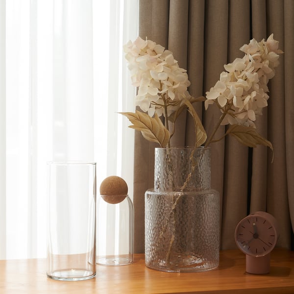 Clear vases and artificial flowers with beige petals and brown stems on a wooden table next to a window with curtains.