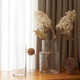 Clear vases and artificial flowers with beige petals and brown stems on a wooden table next to a window with curtains.