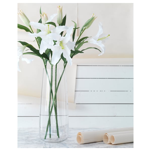 Vase with white lilies on table, white wall, marble countertop, rolled paper.
