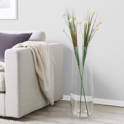 SMYCKA tall clear vase with white base holds artificial yellow flowers, next to grey sofa with blanket.