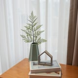 Green vase with plant on books near window.