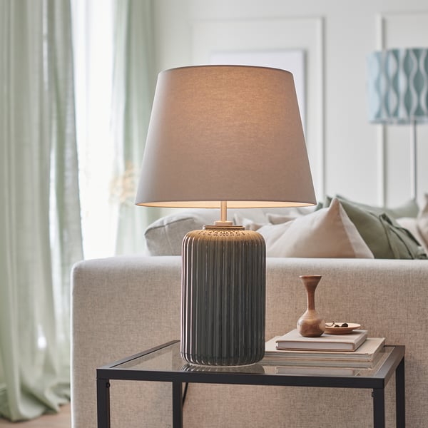 Modern table lamp with ribbed ceramic base, beige shade, on glass table near beige couch.