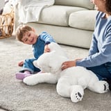 A boy plays with a soft, white SNUTTIG polar bear plush toy on a beige rug. A woman sits nearby, engaging with them.