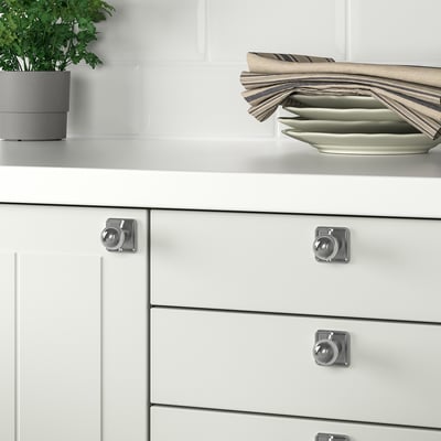 White kitchen drawers with silver knobs and handles, stainless steel plates, and a potted plant.