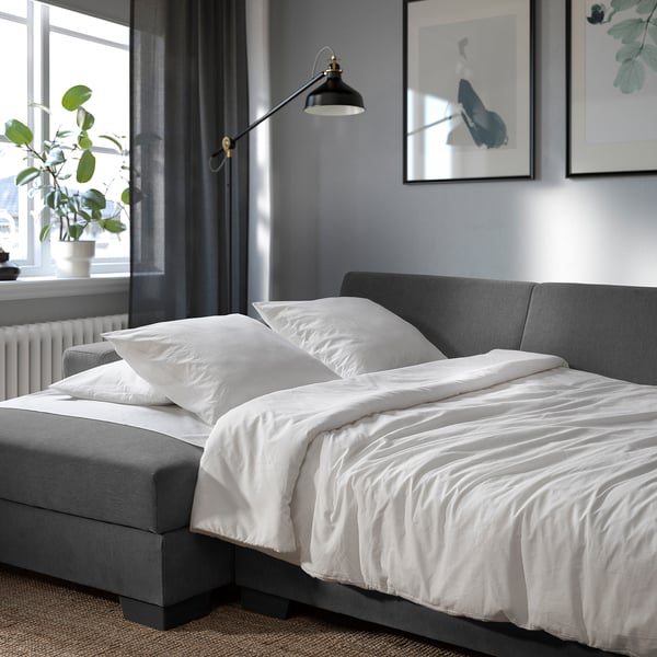 Modern grey sofa bed with white bedding, wall lamp, and plant near window.
