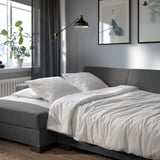 Modern grey sofa bed with white bedding, wall lamp, and plant near window.
