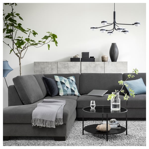 Modern grey l-shaped sofa with blue pillows, glass coffee table, plants, and chandelier in minimalistic living room.