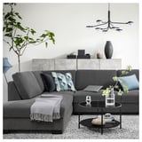 Modern grey l-shaped sofa with blue pillows, glass coffee table, plants, and chandelier in minimalistic living room.