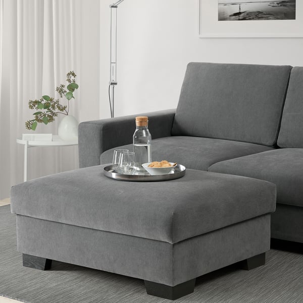 Modern living room with gray l-sofa, footstool used as seat with snacks, white walls, floor lamp, and flowers.