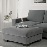 Modern living room with gray l-sofa, footstool used as seat with snacks, white walls, floor lamp, and flowers.