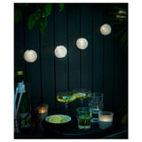 String lights illuminate a patio at night. Glasses, lime slices, and candles rest on a table against a wooden fence.