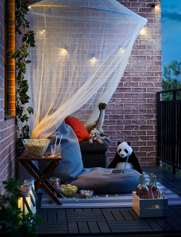 Cozy patio setup with black and blue cushioned chair, white net, string lights, snacks, drinks, and stuffed animals for relaxation.