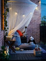 Cozy patio setup with black and blue cushioned chair, white net, string lights, snacks, drinks, and stuffed animals for relaxation.