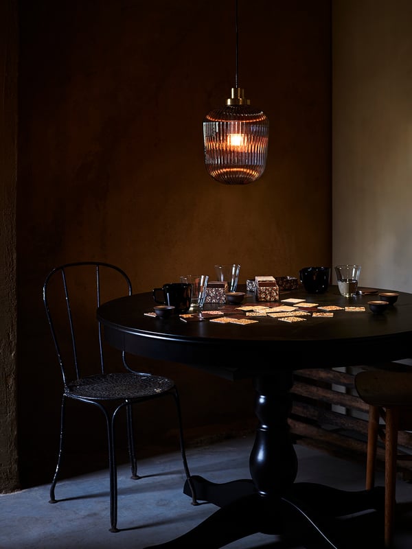Cozy dimly lit room with round dark table, chairs, hanging brass and glass lamp, drinks setup.