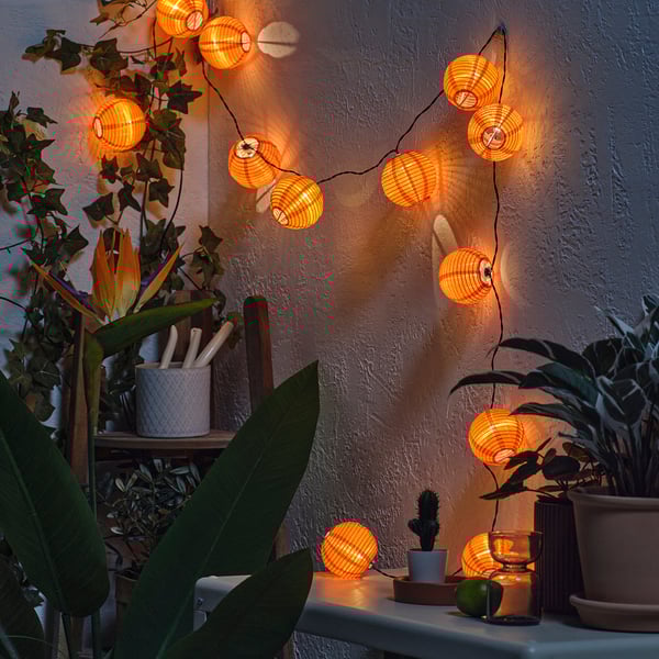 Cozy lit orange lanterns hang across a wall, surrounded by plants and candles, creating a warm ambiance.