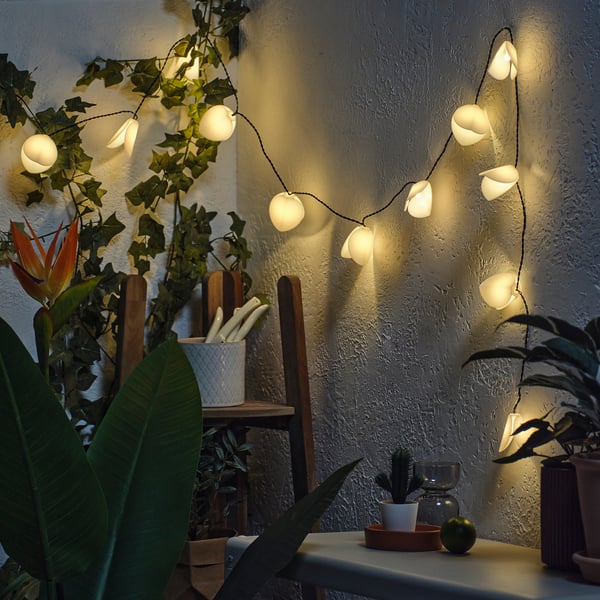 String of solar-powered flower lights illuminating a wall, surrounded by lush green plants in pots.