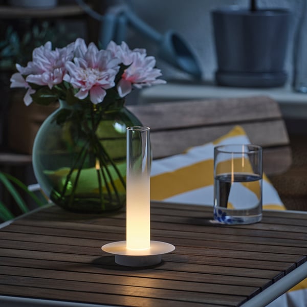 A wooden table displays a lit candle lamp, a vase with pink flowers, and a glass of water at dusk. Ambient lighting provides a cozy setting.
