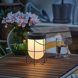 Wooden patio table with a lit solar-powered lantern, a vase of pink flowers, and a glass of water.
