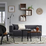 Modern living room with grey sofa, black chair, colorful rug, nesting tables, and wall decor with coffee press.