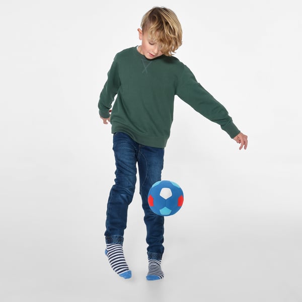 Child balancing colorful ball on knee, wearing green sweater, jeans, and striped socks.