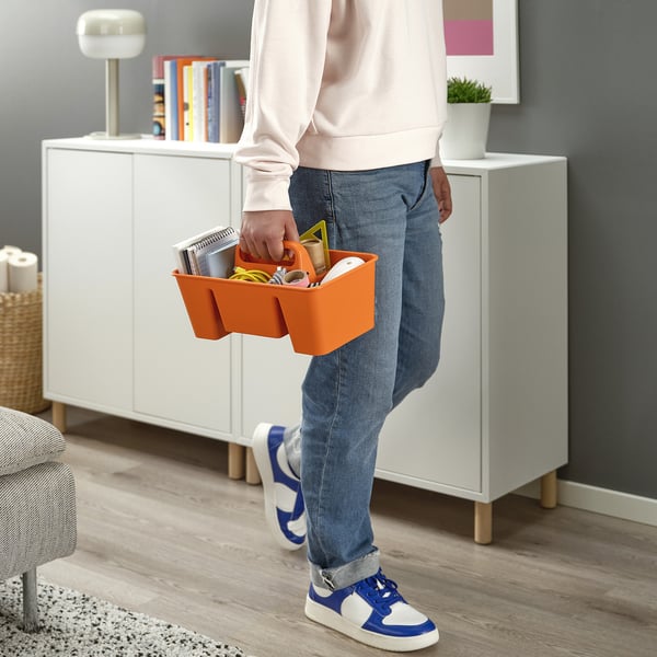 Person holding orange STACKSTOD storage caddy with stationery. White cabinet, lamp, and bookshelf in background.