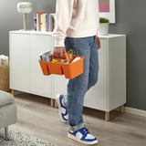 Person holding orange STACKSTOD storage caddy with stationery. White cabinet, lamp, and bookshelf in background.