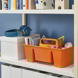 Shelf with blue walls, holding storage bins, headphones, and stationary items. An orange bin stores various small office supplies.