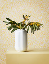 A white ribbed ceramic vase, containing green and yellow flowers, stands on a yellow surface against a yellow patterned wall.