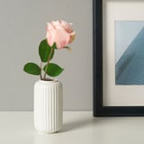 A white ribbed vase holding a single light pink rose with green leaves sits on a white surface next to a black-framed picture.