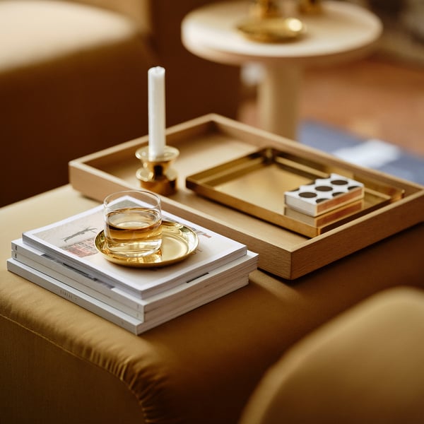 Small golden tray holds items on brown couch, beside stack of books.
