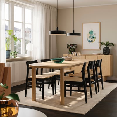 Modern dining room with light wooden table, black chairs, large window, green plant, and minimalist decor.
