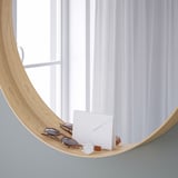 A wooden-framed mirror with a bottom shelf holds sunglasses, a watch, and an envelope. The mirror is positioned by a window with sheer white curtains.