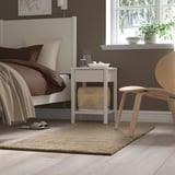 A cozy bedroom with a white rug, brown bed, white table, and wooden chair.