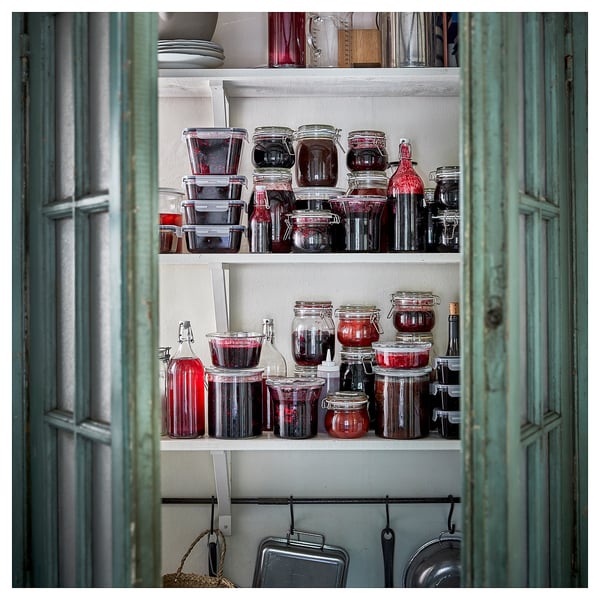 Shelf with various jars, many filled with dark preserve-like contents, in a cozy, rustic room with turquoise green doors.