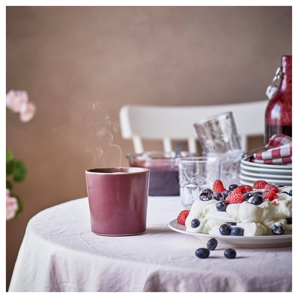 Image a purple ceramic mug with steam, a plate of berries, and glasses. On table with a white tablecloth.