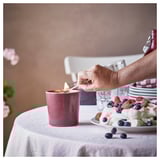 Person lighting pinkish STÖRTSKÖN candle beside fresh fruits and desserts on table.