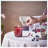A hand lights a red STÖRTSKÖN candle on a table set with a cream berry dessert and a white tablecloth.